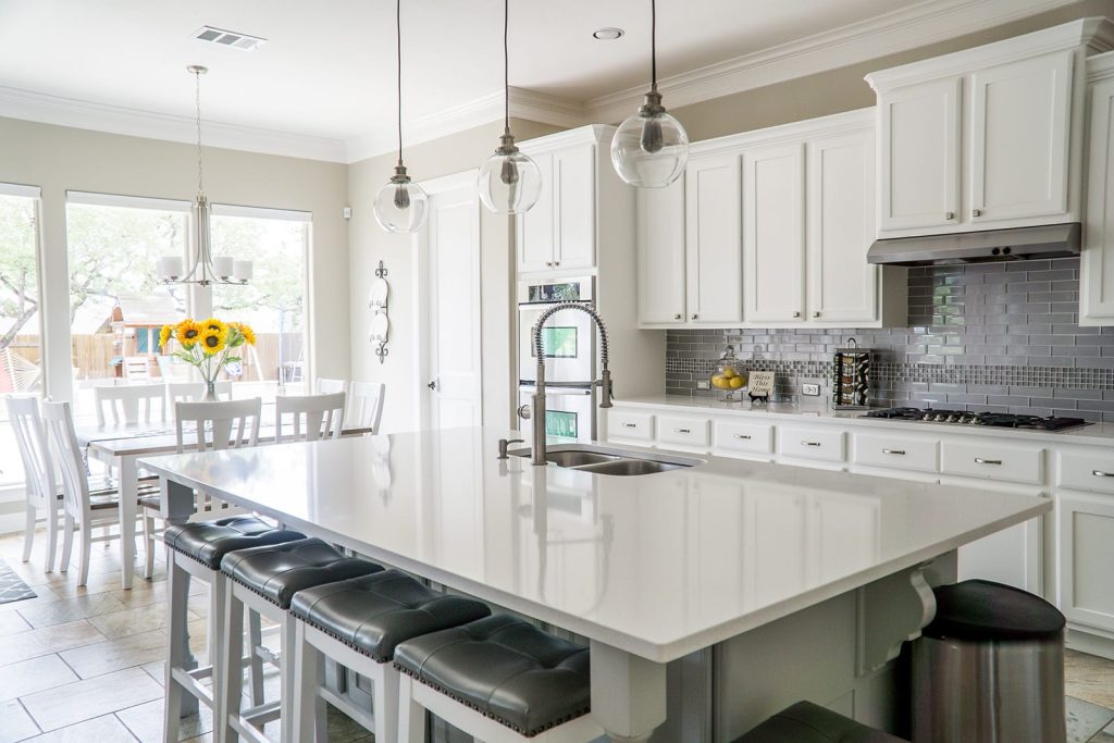 Modern kitchen with white cabinets, gray backsplash, large island with built-in sink, bar stools, stainless steel appliances, and a dining table with sunflowers near large windows.