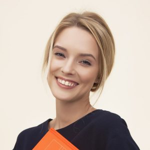 A young woman with blonde hair smiles at the camera while holding an orange book against a light background.