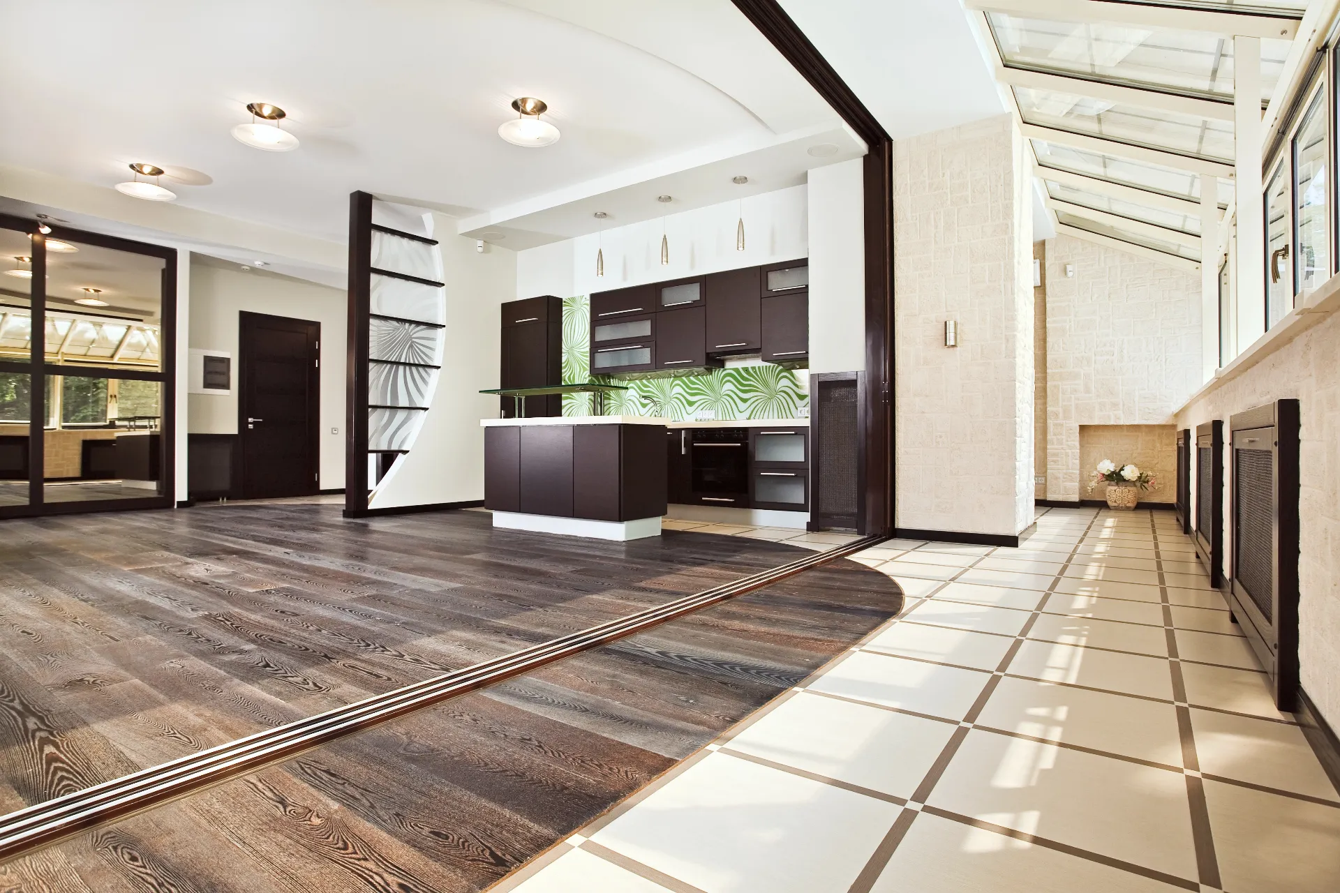 Modern open-plan kitchen and living area with dark wood flooring, white tiled sunroom, and minimalist dark cabinetry with green leaf-patterned backsplash.