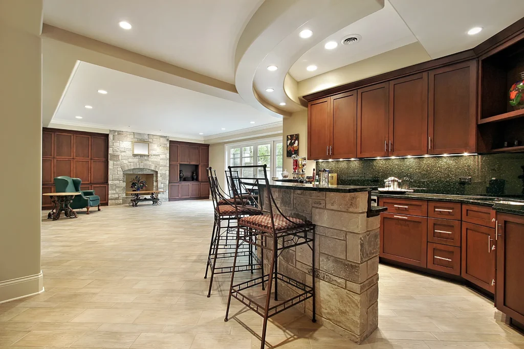Spacious modern kitchen with stone island, dark wood cabinets, bar seating, tile flooring, and an open view to a living area with a fireplace and large windows.
